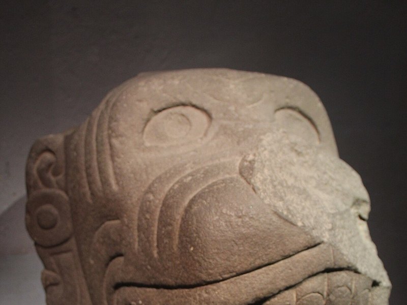 A carved stone head of a god with large round eyes and an open, toothed mouth, in a museum case
