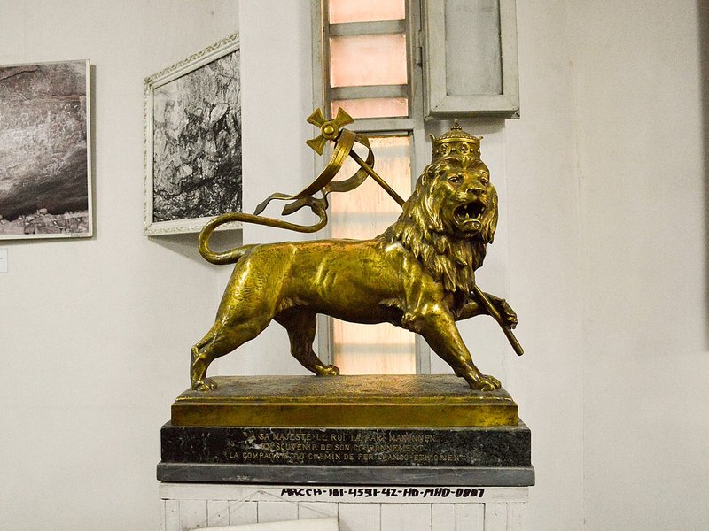 A gold-coloured bronze lion wearing a crown and holding a ribboned cross-topped staff, on a marble base in a museum